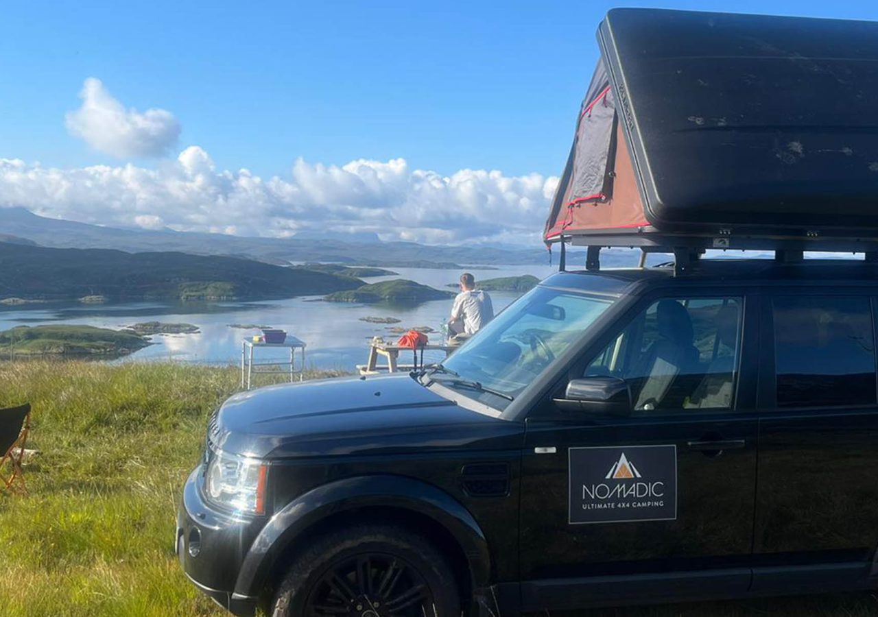 A Nomadic Scotland Land Rover with camping tent on its roof overlooking a a stunning panoramic view of a Scottich Loch