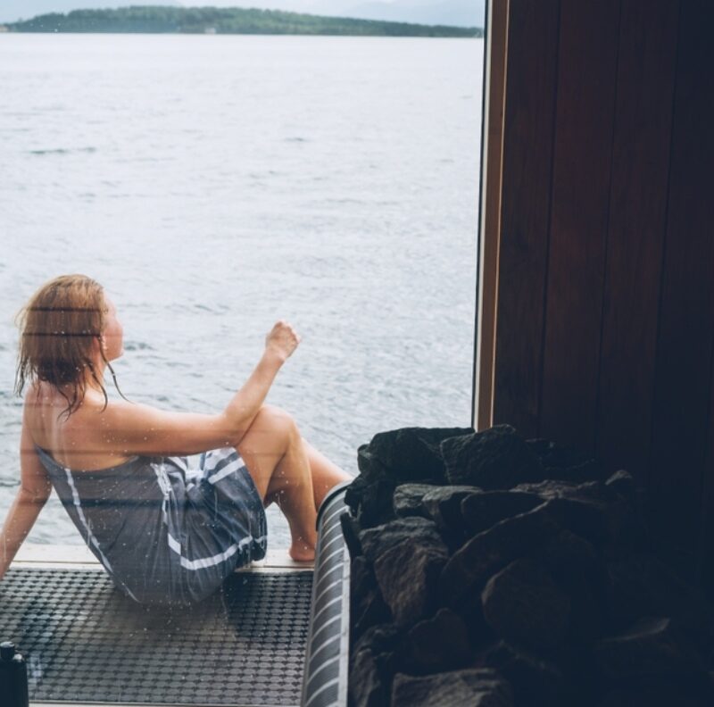 A lady sits out on the decking on an example of a floating sauna on a lake in Scotland.