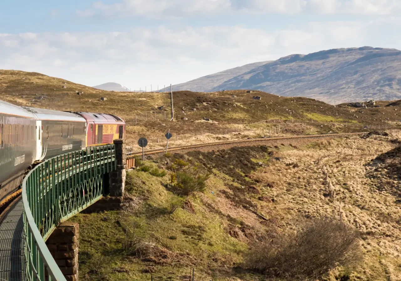 Passenger train crossing a curved bridge through a rugged highland landscape with grassy hills and distant mountains under a partly cloudy sky.