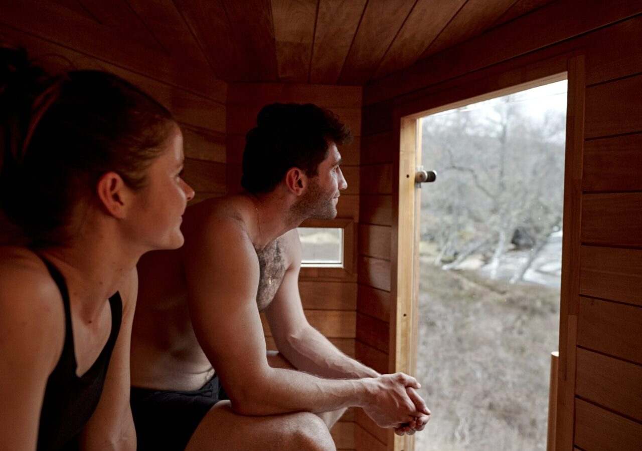 Two people in a wood-lined sauna looking out at the Highland landscape